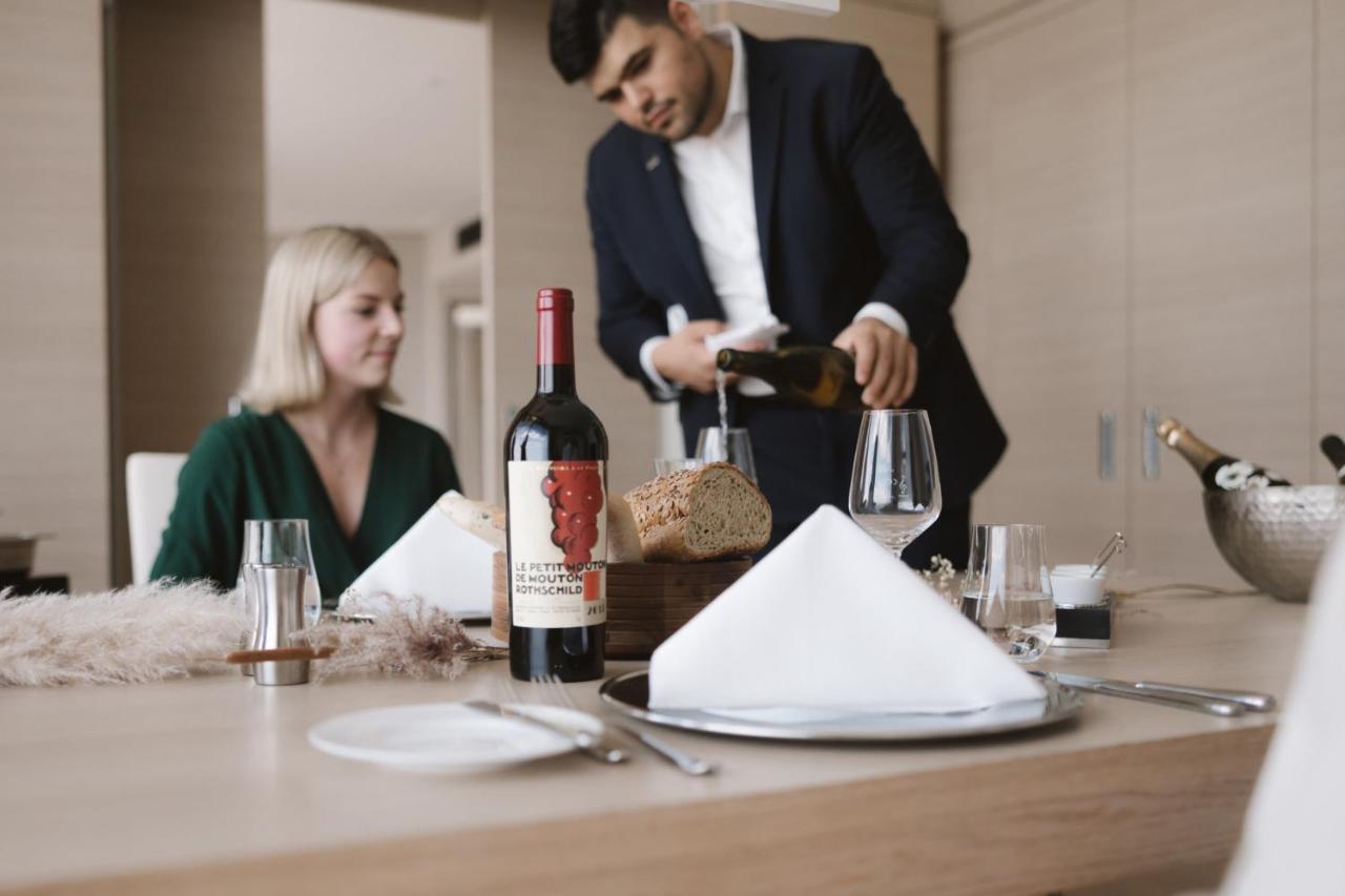 Hotel The Westin Hamburg Elbphilharmonie Exterior foto The photo shows a dining scene featuring a well-dressed man pouring wine into a glass while a woman sits at the table. The table is elegantly set with a folded napkin, wine glasses, and a bottle of red wine placed prominently. There is also a loaf of