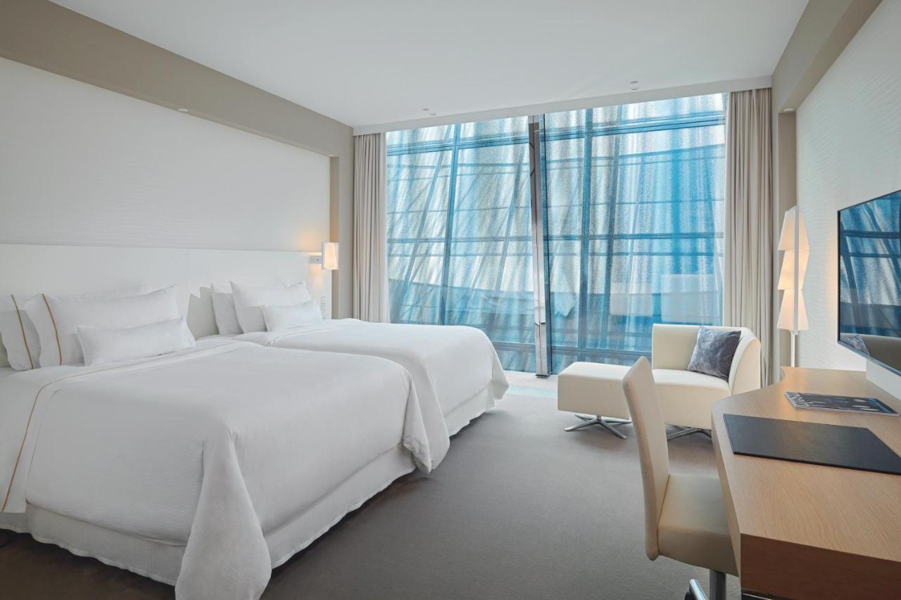 Hotel The Westin Hamburg Elbphilharmonie Exterior foto The photo shows a modern hotel room featuring two white beds with plush bedding. There is a large window with sheer curtains that allows natural light to filter in, offering a view outside. The room has neutral-colored walls and carpeting, creating a