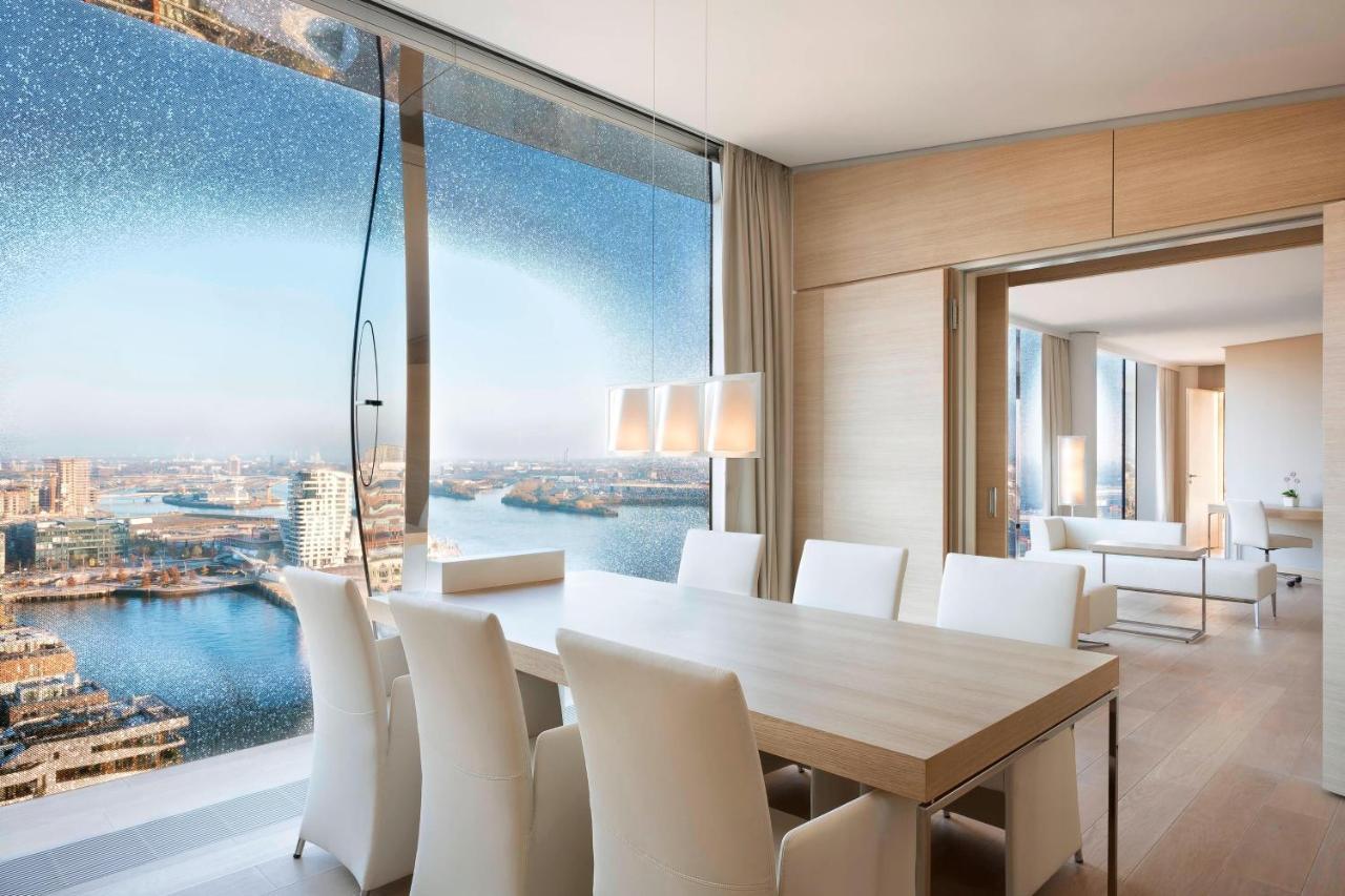 Hotel The Westin Hamburg Elbphilharmonie Exterior foto The photo shows a modern dining area with a large, light-colored table surrounded by white chairs. The space features a panoramic window that offers a stunning view of a body of water and a cityscape. The decor is minimalistic and elegant, with smoot
