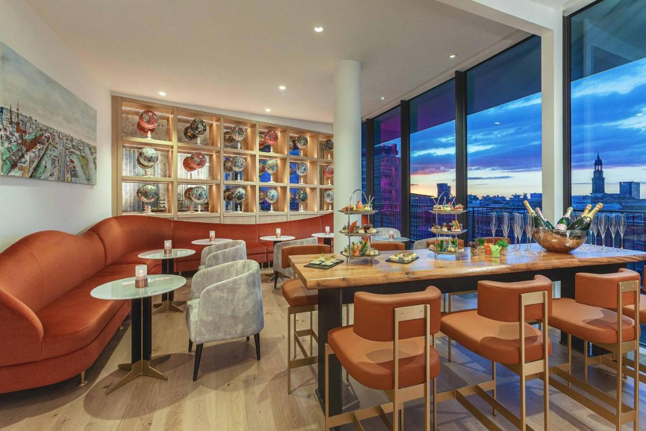 Hotel The Westin Hamburg Elbphilharmonie Exterior foto The photo shows a modern and stylish restaurant or café interior. It features a combination of comfortable seating, including a curved, orange sofa and gray chairs. The tables are round with light-colored tops, and there are several tiered stands dis