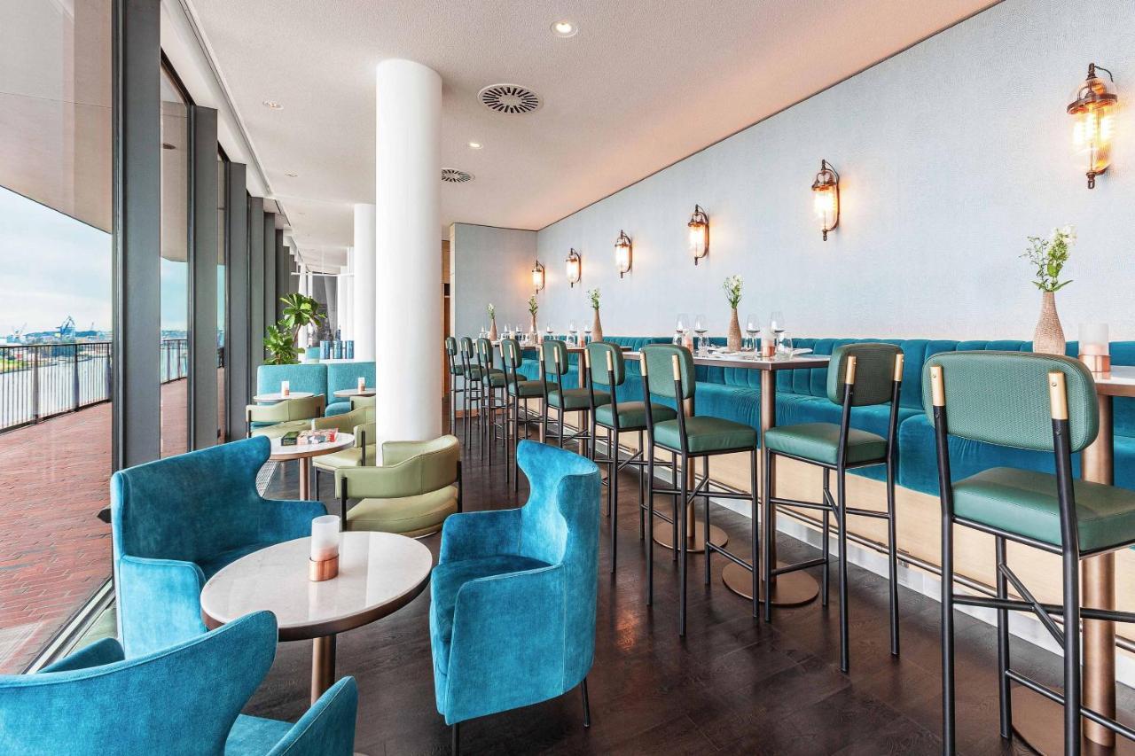 Hotel The Westin Hamburg Elbphilharmonie Exterior foto The photo shows a modern and stylish restaurant or café interior. It features a long row of high-backed bar stools at a counter, complemented by plush seating in the form of rounded chairs and small tables arranged throughout the space. The color sch