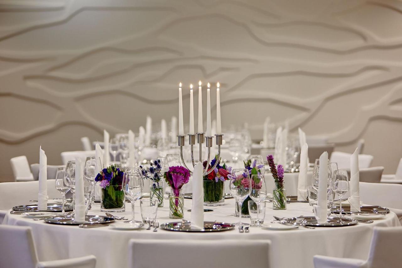 Hotel The Westin Hamburg Elbphilharmonie Exterior foto The photo depicts an elegantly set round dining table. In the center, there is a candelabra with several tall candles, providing a soft, warm light. Surrounding the candelabra are small glass vases filled with colorful flowers, adding a vibrant touch