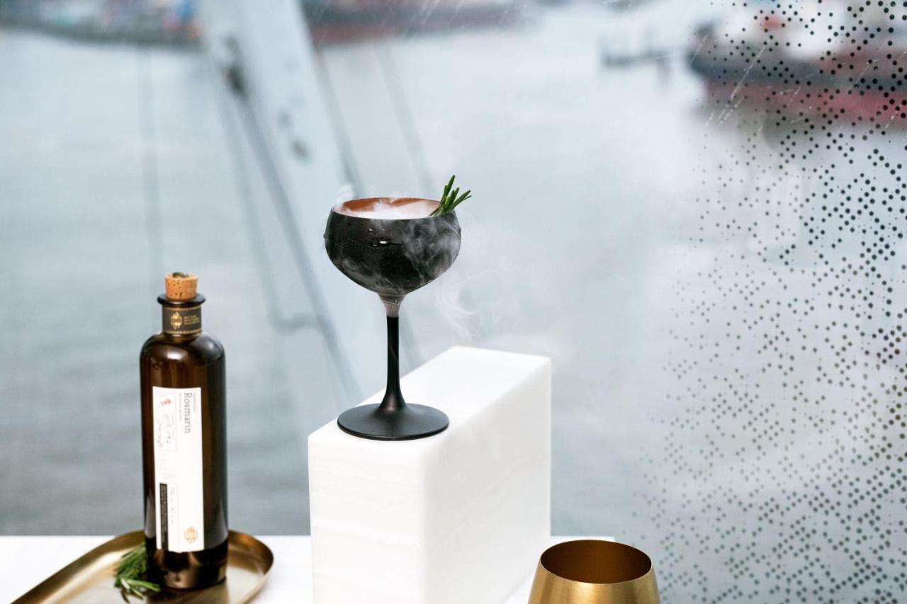 Hotel The Westin Hamburg Elbphilharmonie Exterior foto The photo features a stylish cocktail elegantly presented on a white pedestal. The cocktail is served in a sophisticated black stem glass, topped with a garnish of rosemary. Beside it, there is a dark glass bottle, likely containing a spirit or mixer