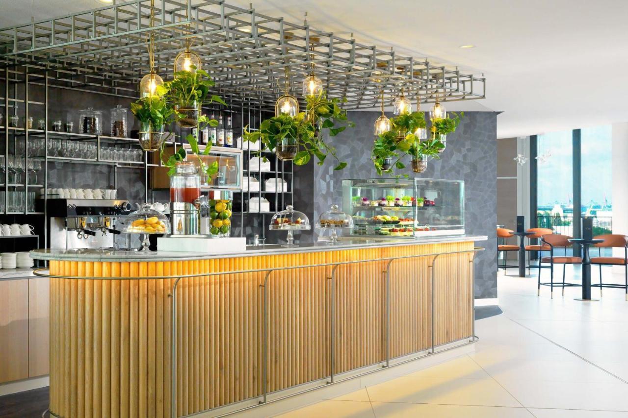 Hotel The Westin Hamburg Elbphilharmonie Exterior foto The photo shows a modern café or bar setting with a sleek wooden counter that features vertical slats. Above the counter, there is a grid-like framework hanging from the ceiling, adorned with various green plants that add a touch of nature to the int