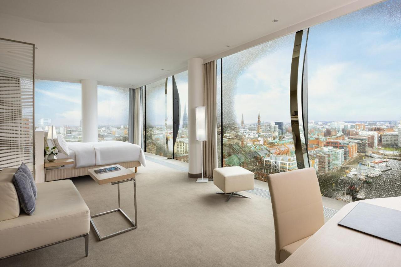 Hotel The Westin Hamburg Elbphilharmonie Exterior foto The photo shows a modern hotel room with large floor-to-ceiling windows that provide a panoramic view of a city landscape. The room features a minimalist design with neutral tones. There's a king-sized bed positioned in the center, a sleek desk and c