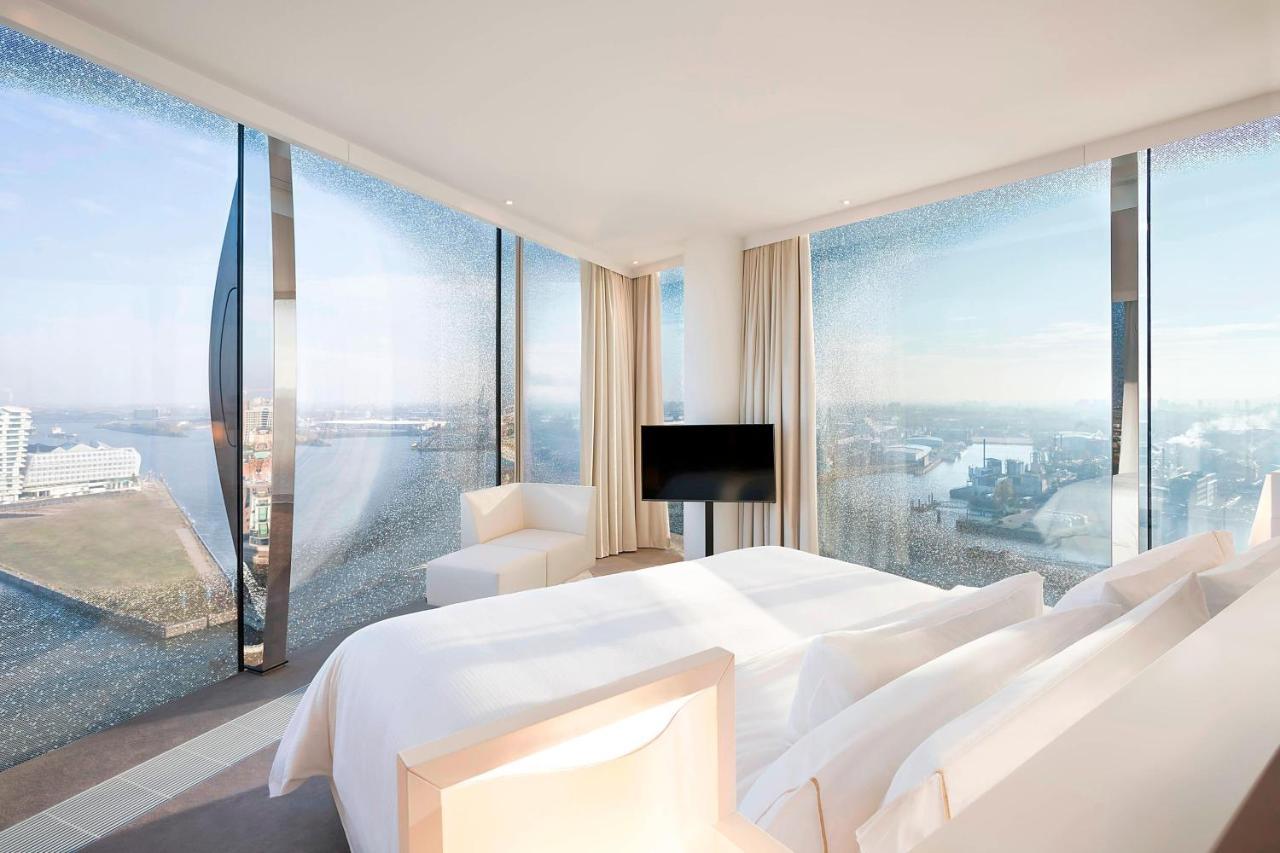 Hotel The Westin Hamburg Elbphilharmonie Exterior foto The photo shows a modern hotel room featuring a large bed with white linens and a minimalist design. It has expansive floor-to-ceiling windows that offer a panoramic view of the landscape outside, likely a city or waterfront setting. The room is deco