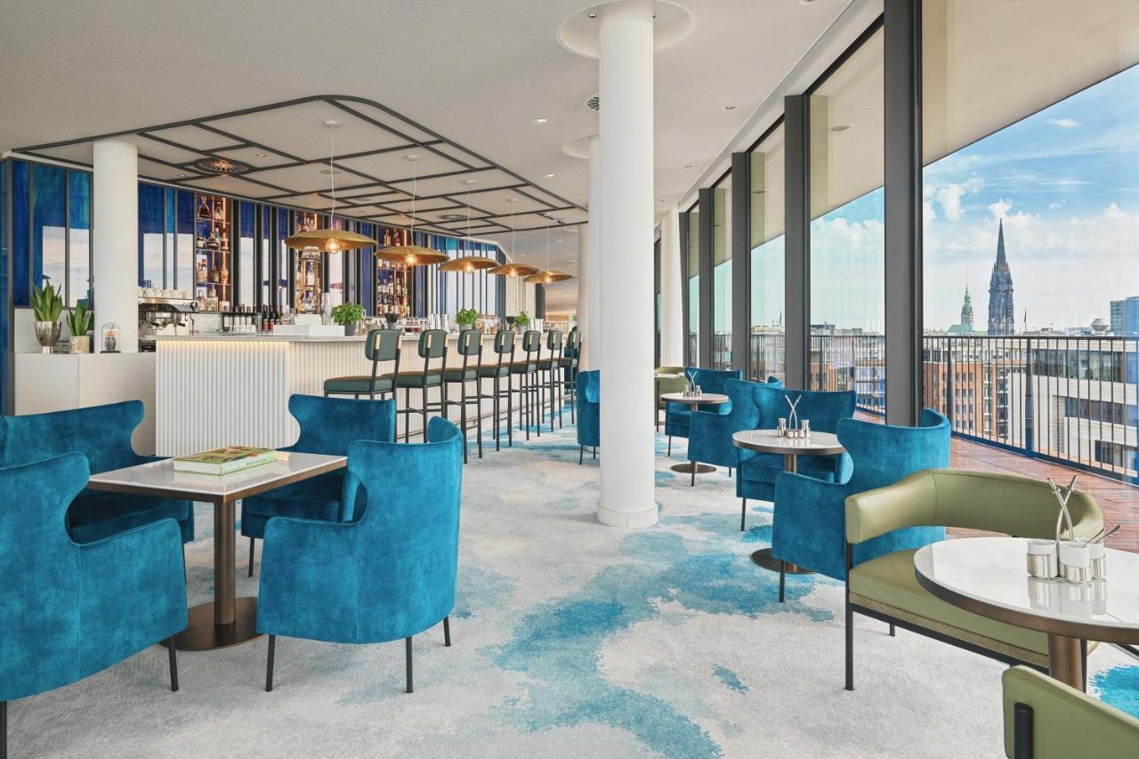 Hotel The Westin Hamburg Elbphilharmonie Exterior foto The image shows a stylish and modern restaurant interior. There are several tables with blue and green chairs, creating a cozy atmosphere. The flooring features a light carpet with abstract patterns. Large windows provide plenty of natural light and 