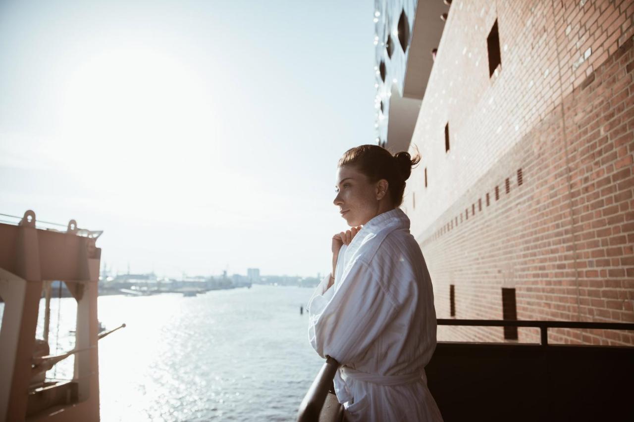 Hotel The Westin Hamburg Elbphilharmonie Exterior foto The photo depicts a person standing on a balcony, looking out over a body of water. The individual is wearing a white bathrobe, and their hair is styled in a bun. The background features a scenic view with sunlight illuminating the scene, creating a 