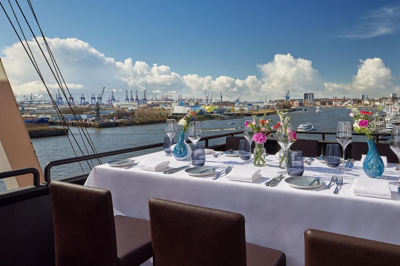 Hotel The Westin Hamburg Elbphilharmonie Exterior foto The photo shows a beautifully set dining table on a rooftop or terrace with a view of a body of water. The table is adorned with a white tablecloth, elegant tableware, and multiple vases filled with colorful flowers. In the background, there are boat
