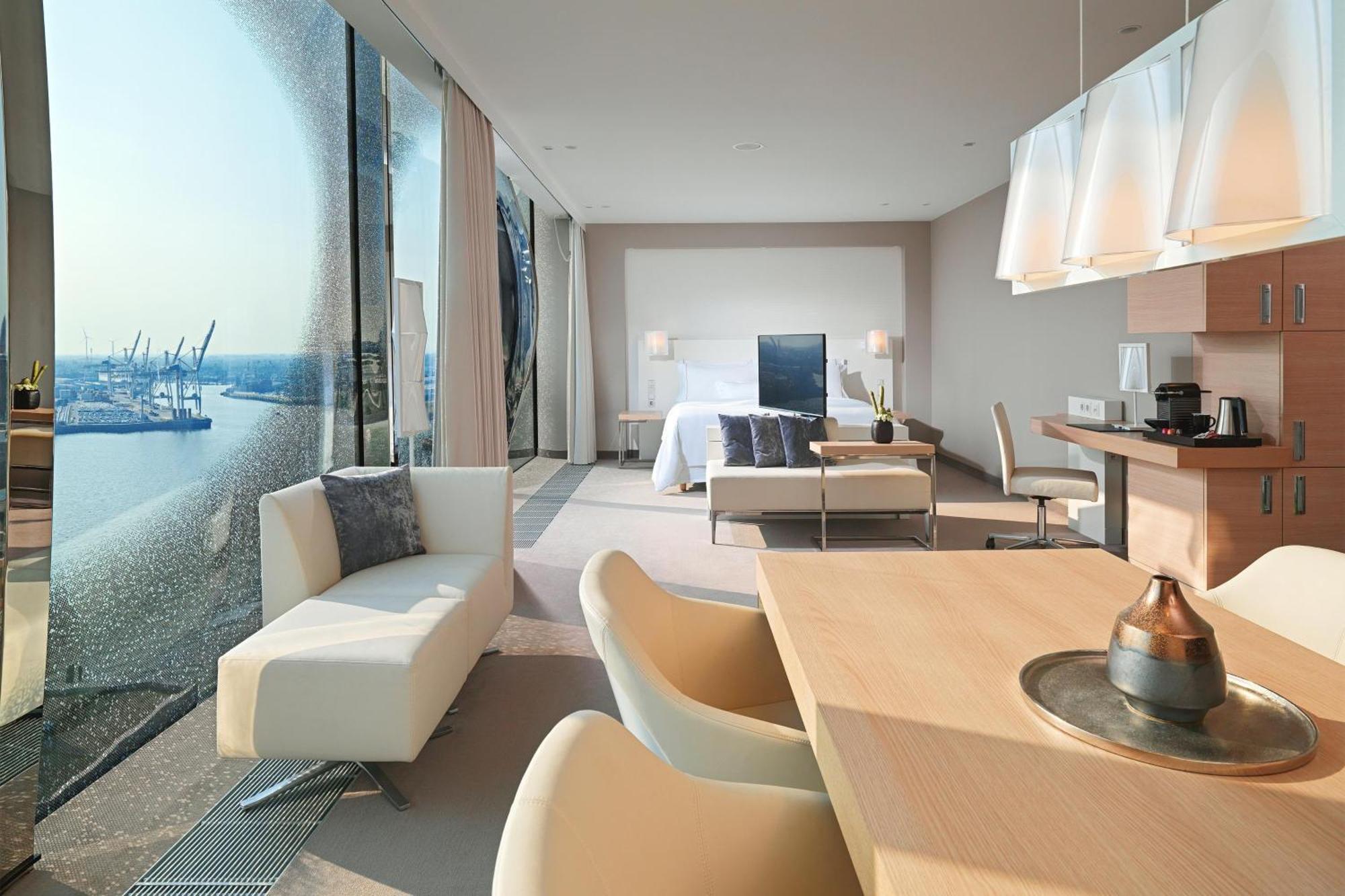 Hotel The Westin Hamburg Elbphilharmonie Exterior foto The photo shows a modern hotel room with a spacious layout. On one side, there are large floor-to-ceiling windows that offer a view of the outside, likely showcasing a waterfront or cityscape. Inside, there's a bed with a minimalist design, accompani