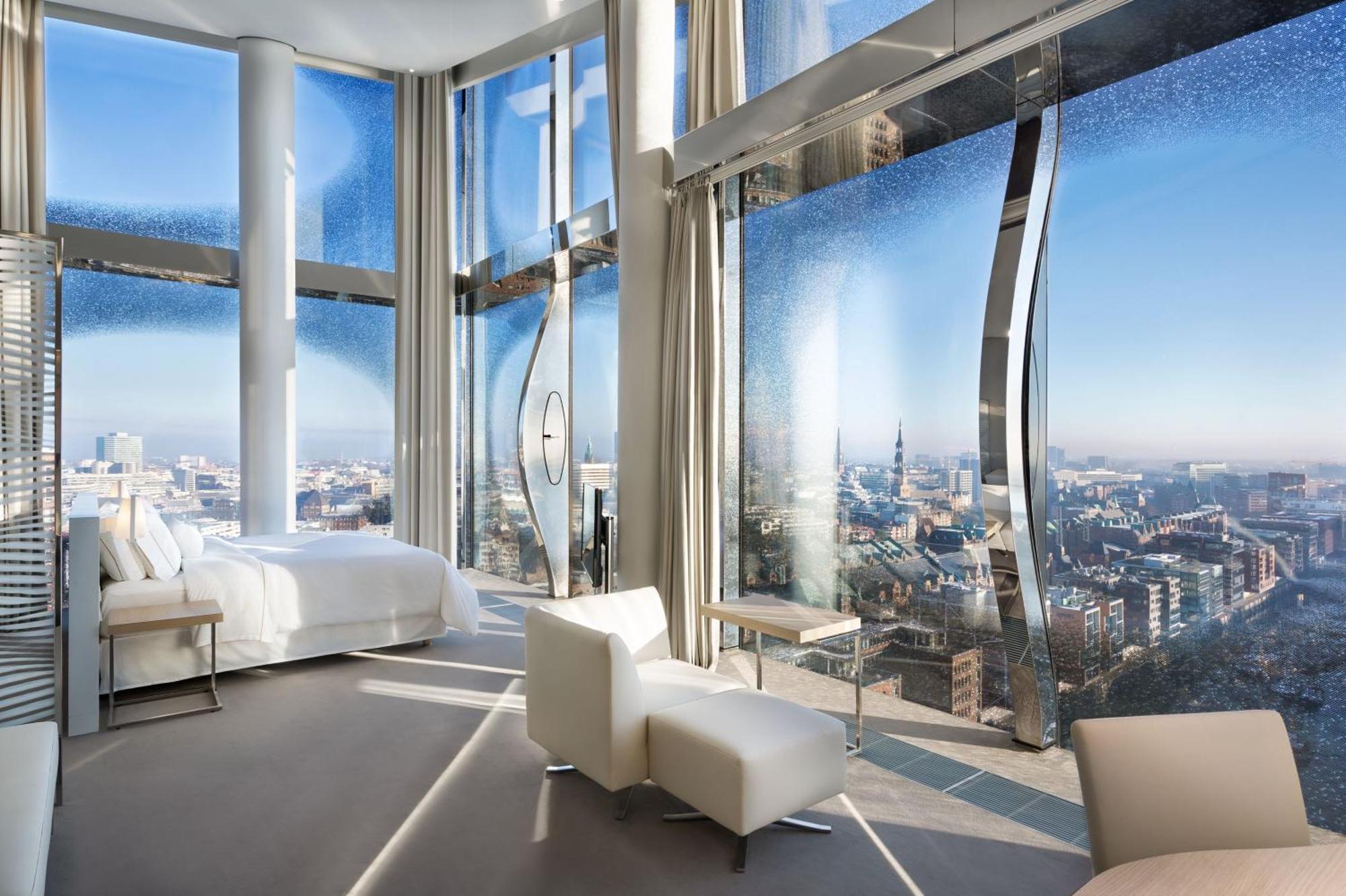 Hotel The Westin Hamburg Elbphilharmonie Exterior foto The photo shows a modern hotel room with a stunning, panoramic view through large, uniquely designed windows. The room features a sleek, minimalist aesthetic with light-colored furnishings, including a white bed and a couple of chairs. The walls are 